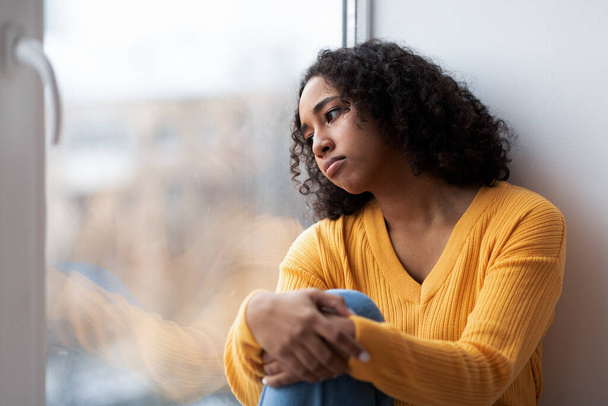 Depressed young African American woman sitting near window, looking outside on street, feeling hopeless at home, copy space. Seasonal mood disorder, mental illness concept - Photo, Image