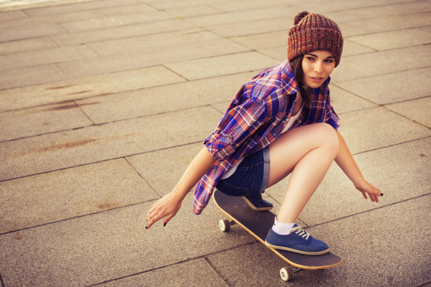 Two Brunette teenage girls friends in hipster outfit (jeans shorts, keds, plaid shirt, hat) with a skateboard at the park outdoors. Copy space - Photo, Image