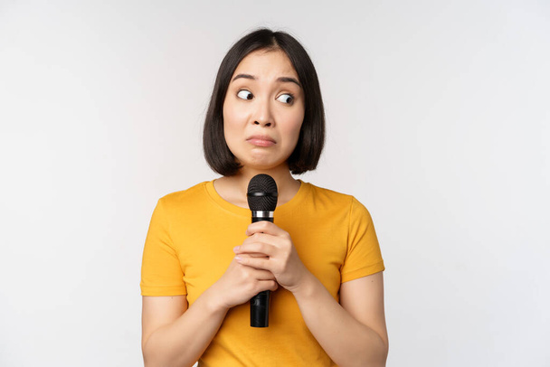 Elinde mikrofon olan mütevazı bir Asyalı kız, halkın önünde konuşmaktan korkuyor, beyaz arka planda duruyor. Boşluğu kopyala - Fotoğraf, Görsel