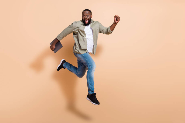 Full body profile photo of impressed young beard guy run hold laptop wear shirt jeans footwear isolated on beige background. - Fotoğraf, Görsel