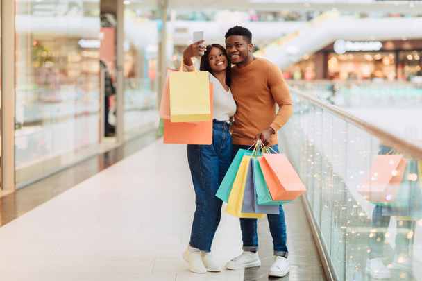 Pár Selfie. Šťastný ležérní Afroameričanka a muž fotí v nákupním centru obchoďáku, usmívá se při pohledu na displej zařízení, drží barevné nákupní tašky, plná délka těla, volný prostor - Fotografie, Obrázek