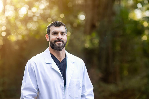 Ένας γιατρός με σκούρα μαλλιά και γενειάδα σε ένα λευκό εργαστηριακό παλτό που στέκεται έξω σε ένα φυσικό πράσινο περιβάλλον με θολό φως. - Φωτογραφία, εικόνα