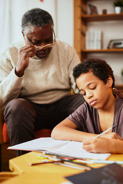 Ein afroamerikanischer Großvater sitzt mit seinem Enkel zu Hause und hilft ihm bei den Hausaufgaben. - Foto, Bild