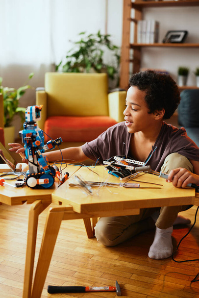 Ein smarter afroamerikanischer Junge hält Lötkolben in der Hand und berührt Tabletten, während er zu Hause einen Roboter baut. Ausbildung in Elektronik. - Foto, Bild
