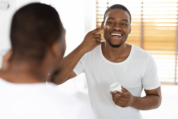 Soins de la peau pour hommes. Heureux bel homme noir appliquant crème hydratante sur le visage tout en se tenant près du miroir dans la salle de bain, homme afro-américain joyeux souriant à sa réflexion, foyer sélectif - Photo, image