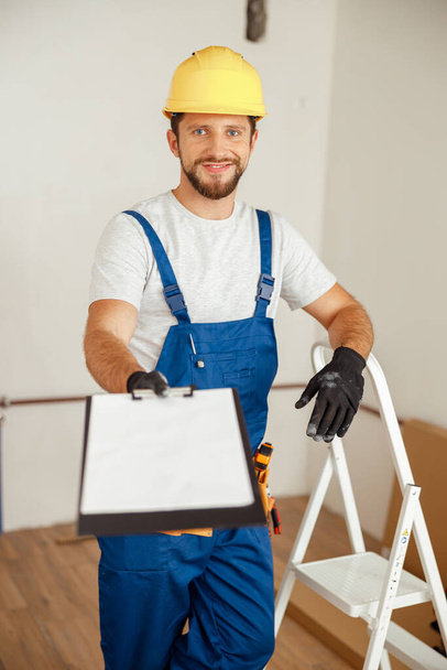 Smiling repairman extending his hand holding clipboard and smiling at camera while standing in new apartment. Manual work, maintenance, repair service concept - Foto, imagen