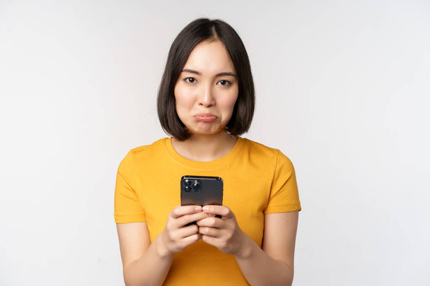 Droevige aziatische vrouw met smartphone, kijken boos met spijt, staan in geel tshirt tegen witte achtergrond. - Foto, afbeelding