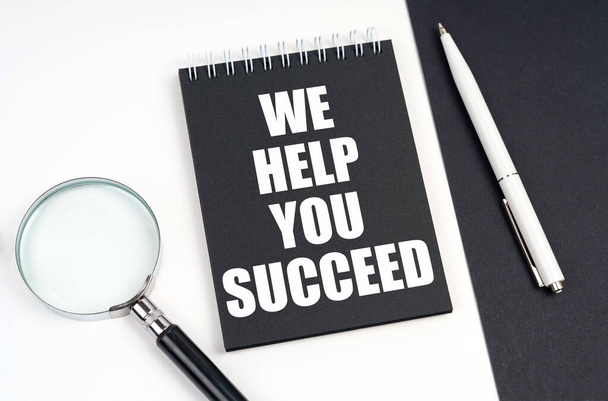 Business and economy concept. On a black and white surface lies a magnifying glass, a pen and a notepad with the inscription - We Help You Succeed - Fotografie, Obrázek