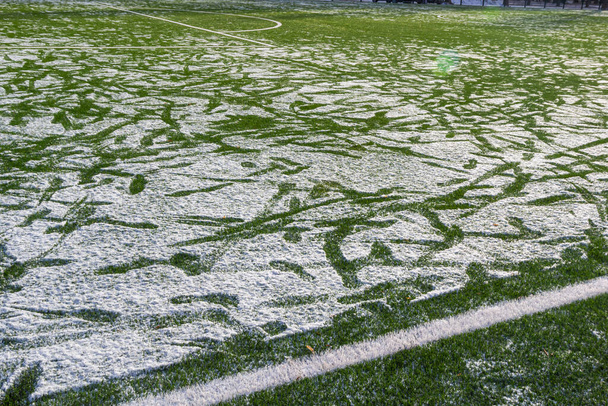 Το γήπεδο ποδοσφαίρου με τεχνητό πράσινο γρασίδι καλύπτεται με ένα ελαφρύ στρώμα χιονιού. Νωρίς άνοιξη. Πράσινο γρασίδι στο γήπεδο ποδοσφαίρου είναι ορατό κάτω από το χιόνι. Ερασιτεχνικό γήπεδο ποδοσφαίρου. - Φωτογραφία, εικόνα