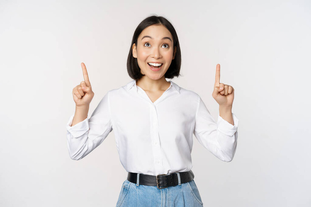 Enthousiaste Aziatische zakenvrouw die wijst, opkijkt met een vrolijk glimlachend gezicht, bedrijfslogo of spandoek toont, over een witte achtergrond staat. - Foto, afbeelding