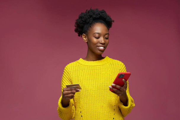 Mobile banking, e-commerce. Smiling african american girl customer hold mobile phone credit bank card make online purchase, payment in e-bank app. High quality photo - Zdjęcie, obraz