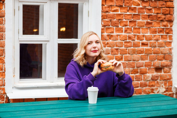 Blonde woman having lunch, coffee break, preparing to eat cheeseburger and drink coffee outdoors - Foto, Bild