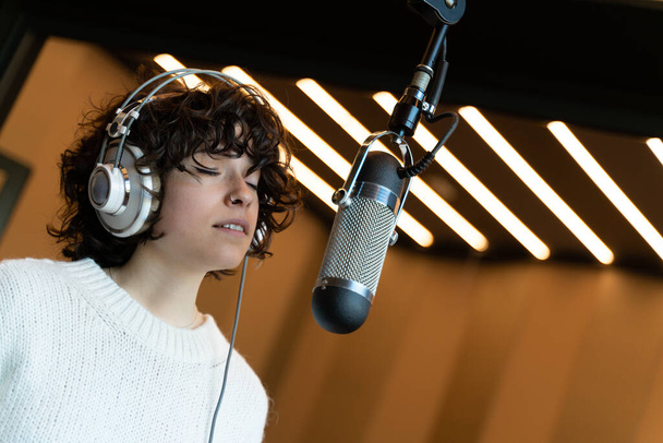 Молода кучерява співачка записує пісню в реальній студії з професійним мікрофоном. Вона в білому светрі
. - Фото, зображення