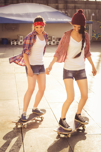 Two Brunette teenage girls friends in hipster outfit (jeans shorts, keds, plaid shirt, hat) with a skateboard at the park outdoors. - Φωτογραφία, εικόνα