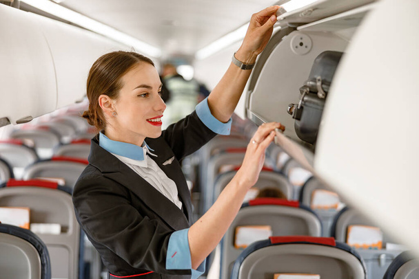 Весела жінка бортпровідник дивиться на проїзну валізу в багажному відсіку і посміхається, стоячи в салоні пасажира літака
 - Фото, зображення