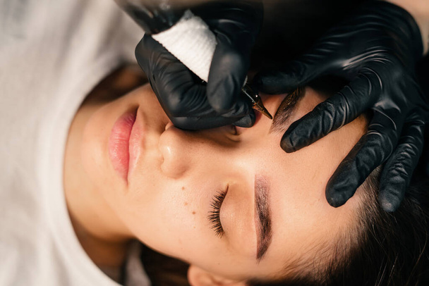 美容師の手は永久的な眉メイクを行います。皮膚への最小限の外傷。マニピュレータハンドルと針付きの特殊ノズルを使用して眉マイクロブレードを実行します。美容師のスキルレベル - 写真・画像