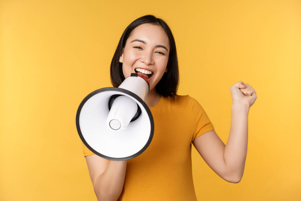 Lächelnde asiatische Frau, die mit Megafon vor gelbem Hintergrund steht und etliches Werbeprodukt ankündigt. - Foto, Bild