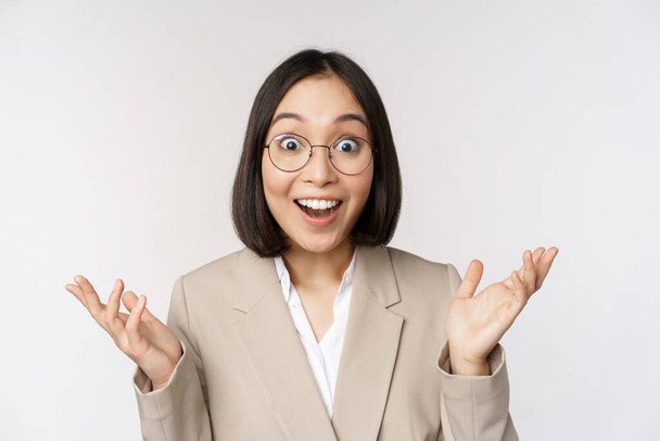 Entuzjastyczna azjatycka sprzedawczyni w okularach, uśmiechnięta i śmiejąca się, wyglądająca na zachwyconą kamerą, stojąca w beżowym garniturze na białym tle. - Zdjęcie, obraz