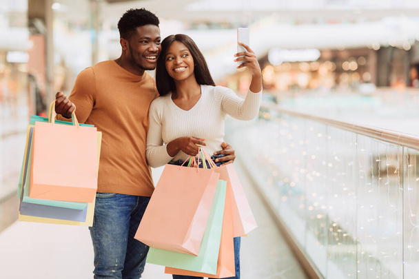 Je m'amuse. Heureux couple afro-américain prenant selfie après avoir fait du shopping dans le centre commercial de la ville, debout avec de nombreux sacs à provisions, souriant à la caméra gadget, tenant en main le smartphone, espace de copie gratuit - Photo, image