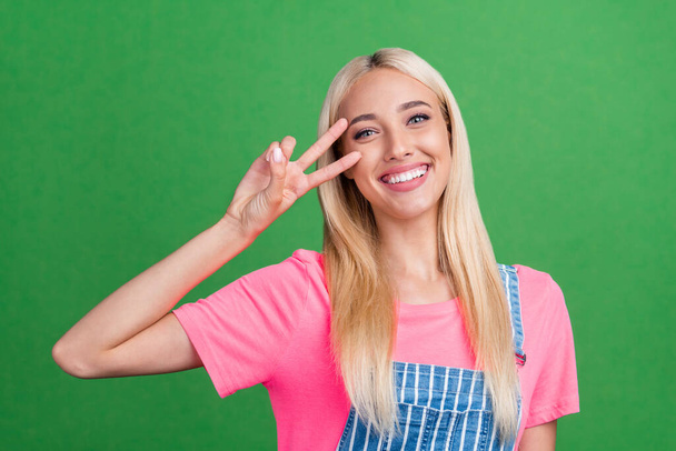 Photo of cheerful friendly lady show v-sign symbol cover eye wear striped overall isolated green color background. - Photo, image