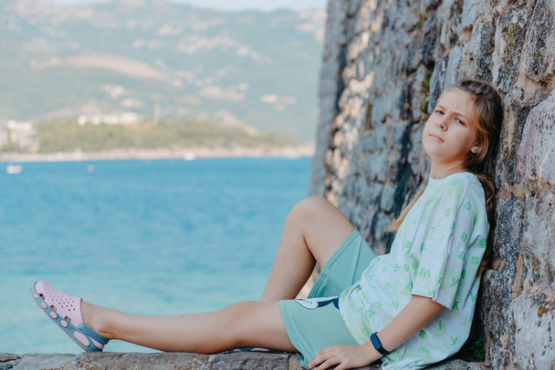 Beautiful girl sitting on a stone wall, in background is the blue sea, Budva, Montenegro. - 写真・画像