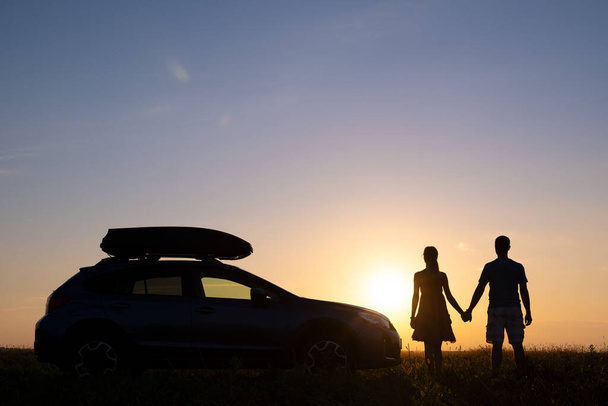 Pareja feliz de pie cerca de su coche al atardecer. Joven hombre y mujer disfrutando del tiempo juntos viajando en vehículo - Foto, Imagen