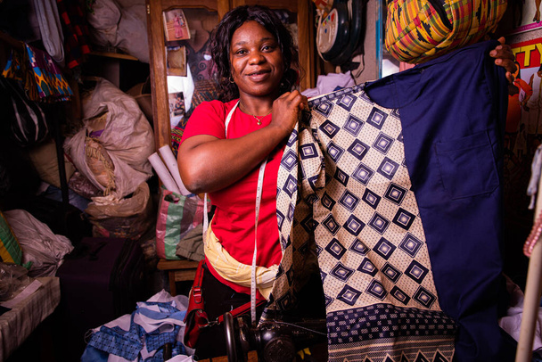 Μια χαρούμενη Αφρικανή μοδίστρα επιδεικνύει το φόρεμα που μόλις τελείωσε το ράψιμο. Τυπικό αφρικανικό κοστούμι για τους άνδρες μόλις έκανε - Φωτογραφία, εικόνα