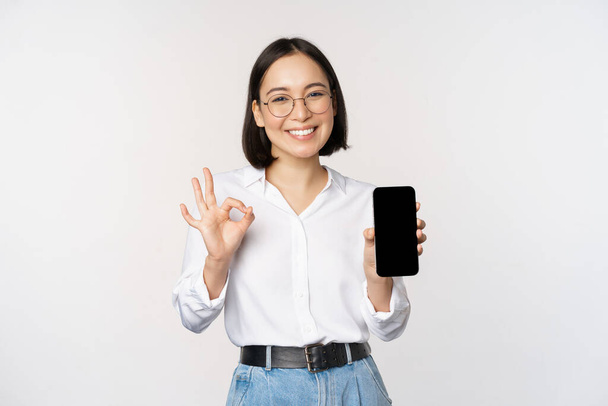 Imagen de mujer de negocios asiática, mostrando la pantalla del teléfono inteligente, interfaz de la aplicación y signo de ok, recomendando la aplicación en el teléfono móvil, de pie sobre fondo blanco. - Foto, Imagen