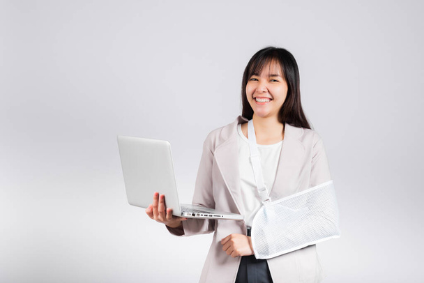 Asian business woman confident smiling broken arm after accident and wear arm splint for treatment but still have to work she hold laptop computer studio isolated on white background,copy space - Φωτογραφία, εικόνα