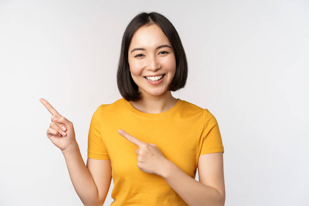 retrato de sorrindo asiático morena menina no amarelo tshirt, apontando dedos esquerda, mostrando cópia espaço, promo negócio, demonstrando banner, pé sobre branco fundo. - Foto, Imagem