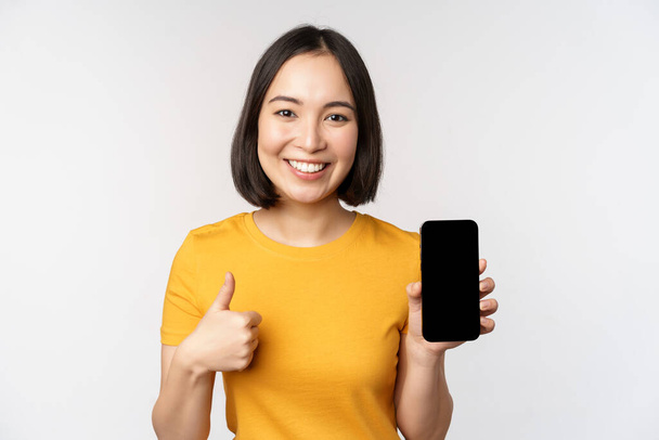 Technology and people concept. Smiling young woman showing thumb up and smartphone screen, mobile phone app interface, standing against white background. - Foto, afbeelding