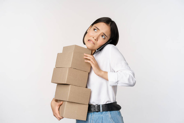 Jeune femme d'affaires asiatique répondre à l'appel téléphonique, parler sur mobile tout en portant pile de boîtes avec des commandes, debout sur fond blanc. - Photo, image