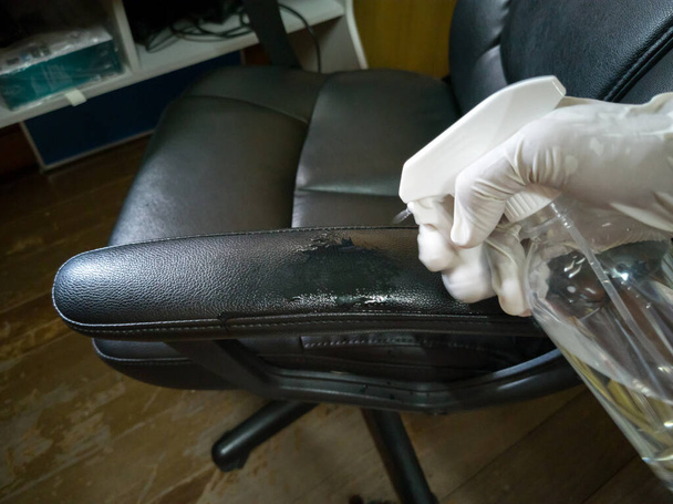 Άποψη υψηλής γωνίας, καθαρισμός μαύρης καρέκλας εργασίας (λάθος και επικίνδυνη μέθοδος), ψεκασμός με οινόπνευμα, ψεκασμός των μπράτσων, λευκά γάντια χέρια. - Φωτογραφία, εικόνα