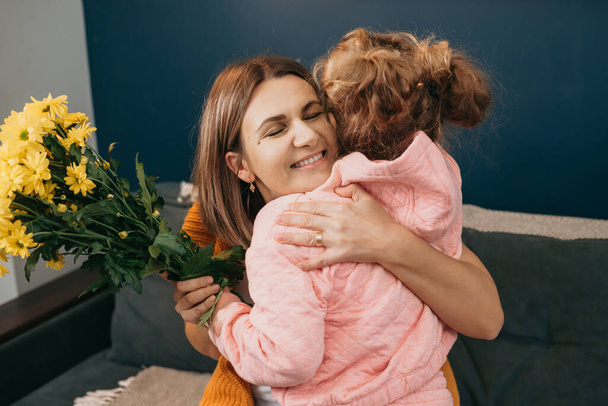 Счастливая мать обнимает свою маленькую девочку, которая поздравила ее с букетом цветов на день рождения матери. Концепция празднования дня рождения. Семейный день. Семья - Фото, изображение