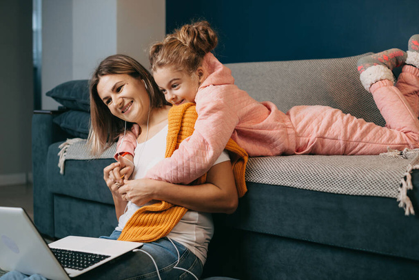 The girl is lying on the couch and hugging her mother who is trying to focus on the work, which she does from a distance due to the pandemic. Happy family - Photo, Image