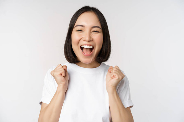 Portret van enthousiaste Aziatische vrouw die wint, viert en triomfeert, handen opsteekt, doel of succes bereikt, over een witte achtergrond staat. - Foto, afbeelding