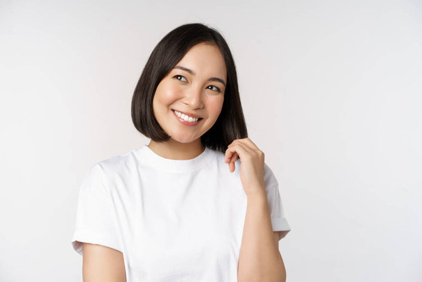 Portrait of cute coquettish woman laughing and smiling, looking aside thoughtful, thinking or imaging smth, standing in white t-shirt over studio background. - Photo, Image