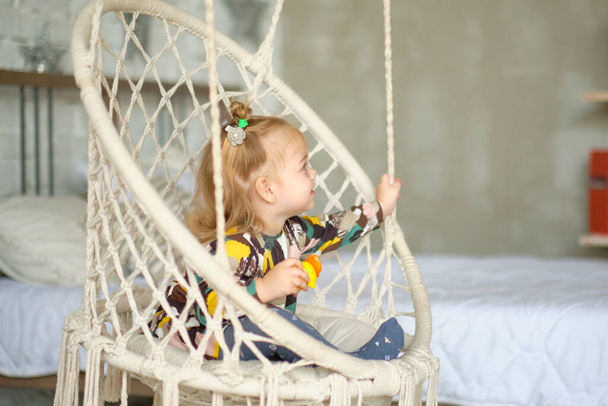 A cute little girl with pigtails on her head is swinging at home in a hanging swing - Foto, afbeelding