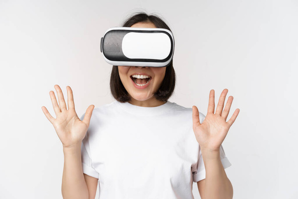 Szczęśliwa azjatka za pomocą zestawu słuchawkowego VR, machając podniesionymi rękami i śmiejąc się, używając okularów wirtualnej rzeczywistości, stojąc nad białym tłem. - Zdjęcie, obraz