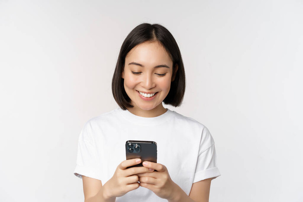 Portrait of smiling asian woman using mobile phone, chatting, texting message, standing in tshirt over white background. Copy space - Photo, Image