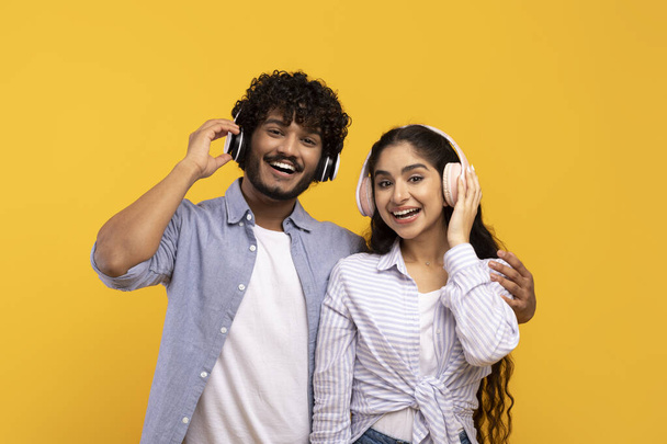 Szczęśliwa indyjska para w słuchawkach słuchająca muzyki i przytulająca się do żółtego studyjnego tła. Podekscytowany mężczyzna i kobieta cieszący się ulubionymi ścieżkami dźwiękowymi - Zdjęcie, obraz