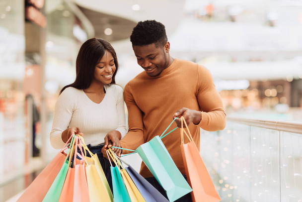 Портрет счастливой улыбающейся афро-американской пары, держащей и смотрящей внутрь красочных торговых мешков, довольной своей новой одеждой или подарком, наслаждающейся совместными покупками, стоящими в центре торгового центра - Фото, изображение