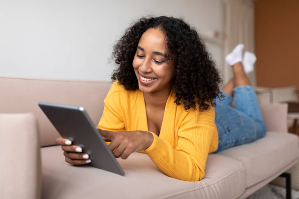 Расслабленная молодая черная женщина лежит на диване и пользуется планшетным ПК, чатится в Интернете или проверяет социальные сети. Веселая афроамериканка с сенсорным блокнотом, охлаждающаяся на диване дома - Фото, изображение