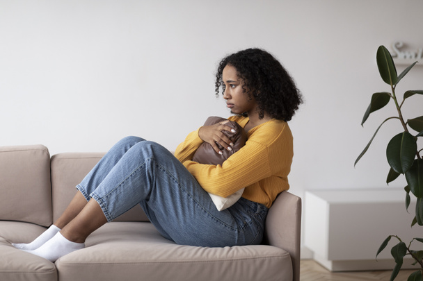 Unhappy young black woman suffering from depression, hugging pillow, sitting on couch at home. Upset African American lady feeling hopeless, having mental illness or post traumatic stress disorder - Foto, afbeelding