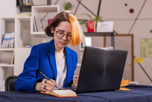Νεαρό σύγχρονο κορίτσι με πολύχρωμα μαλλιά σε μπλε κοστούμι επιχειρήσεων κάθεται στο τραπέζι με φορητό υπολογιστή. Κάνει σημειώσεις σε ένα πορτοκαλί σημειωματάριο. - Φωτογραφία, εικόνα