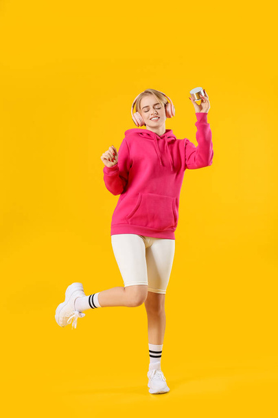 Happy young woman with headphones holding wireless portable speaker and dancing on color background - Foto, afbeelding