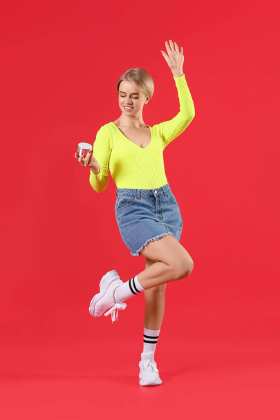 Young woman with wireless portable speaker dancing on red background - 写真・画像