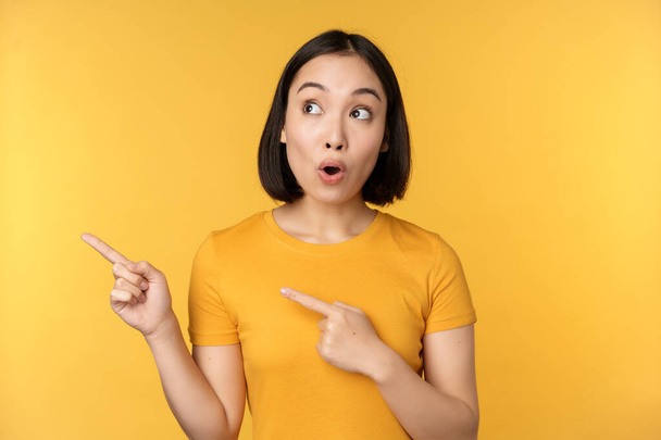 Ritratto di donna asiatica intrigata, guardando e indicando il dito sinistro alla pubblicità, mostrando smth interessante, in piedi su sfondo giallo. - Foto, immagini