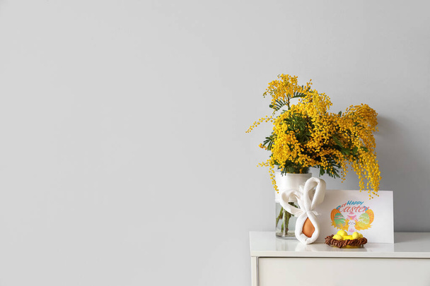 Cartão de saudação com texto HAPPY EASTER, flores mimosa em vaso e ovos na prateleira perto da parede de luz - Foto, Imagem