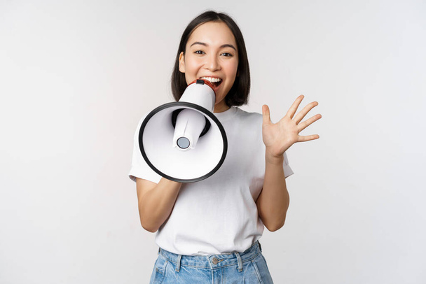Щаслива азіатська жінка кричить на мегафон, роблячи оголошення, рекламуючи щось, стоїть на білому тлі
. - Фото, зображення